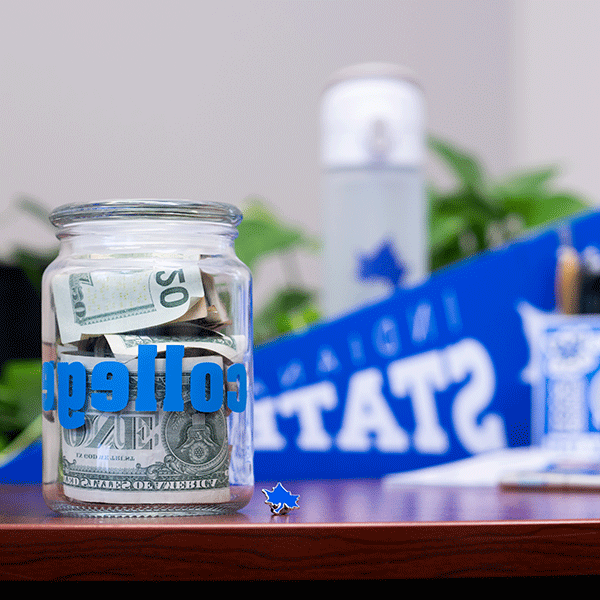 A clear glass jar with ‘COLLEGE’ printed on it in blue letters sits on a desk. It is filed with bills of various denominations. A folded $20 is visible, as is the back of a $1 bill. A blue Indiana State pennant is visible but out of focus in the background, along with a white and gray thermos with a blue Sycamore leaf on it, a sheet of stickers, and a few other items. Greenery and a white wall are visible in the background.